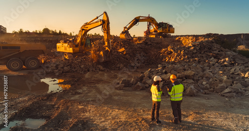 Aerial Drone Shot Of Construction Site On Sunny Evening: Industrial Excavators Digging Rocks And Loading Into A Truck. Civil Engineer And Inspector Observing Process, Discussing Real Estate Project