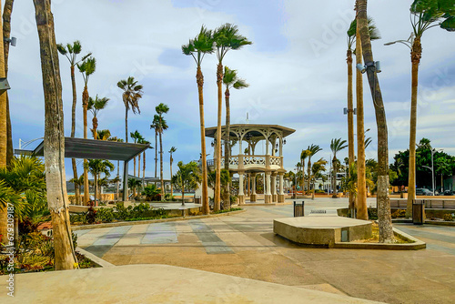 Panoramic view of promenade or malecon with its kiosk, palm trees, concrete benches, sky covered with clouds in background, cloudy spring day in La Paz, Baja California Sur Mexico