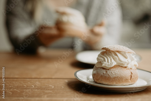 Focus on traditional Swedish pastry, semla, on a plate at wooden table. In the background, out of focus, a person is about to eat pastry. Photo taken in Sweden.