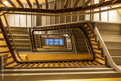 View to top of highlighted high and old square spiral staircase with decorative wooden railings and yellow ceilings with glass skylight on top