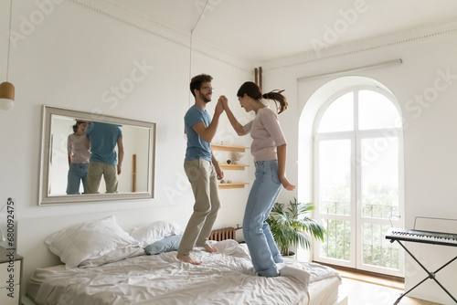 Young millennial carefree couple spend free time at home jumping on bed in cozy bedroom, have fun, enjoy family weekend at modern apartment, celebrating purchase of new house. Date, relationship, mood