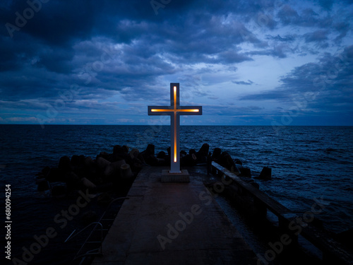 A large Christian cross stands at the edge of a pier against a dramatic sky and sea, seen from above