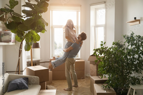 Happy husband lifts his young beautiful wife, laughing, swirling, enjoy move-in day to new, first house, big carton boxes nearby in unfurnished sunny living room. Bank loan for young family, new life