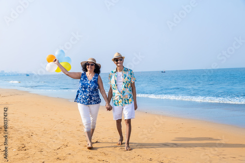 Retired Senior indian couple with colorful balloon in hands walking together at beach. Enjoying vacations holidays at beach. Love, romantic, bonding, relationship, fun, Copy space.