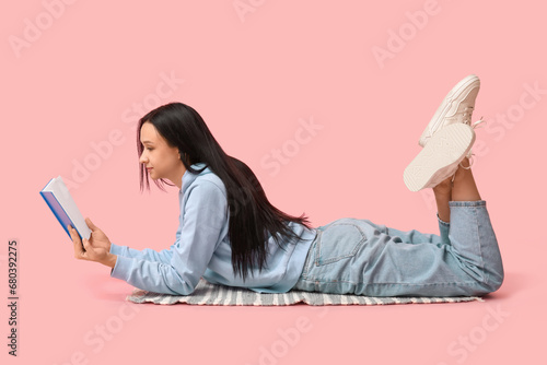 Beautiful young woman lying on pink background and reading book