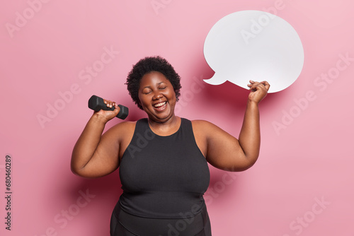 Obese dark skinned African woman dressed in black tracksuit lifts dumbbell has aerobics or gymnastics smiles positively holds blank speech bubble for your advertisement isolated over pink background
