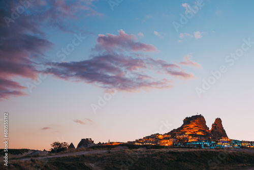 The fading light of day paints the skies over Cappadocia with strokes of pink and blue, while the town below basks in the warm glow of evening lights, nestled among ancient rock formations.