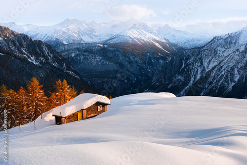 Lonely wooden house on snowy meadow surrounded by orange larch forest in Italian Alps. Monte Bianco mountain range on background. Location place: Courmayeur, northern Italy. Landscape photography