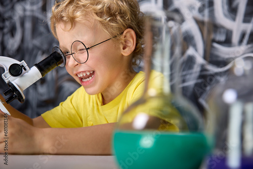 Young scientist of five years, squinting, attentively looks with one eye into the eyepiece of microscope, holding a tripod with his hand. Happy Caucasian boy in a yellow T-shirt studies a microscope