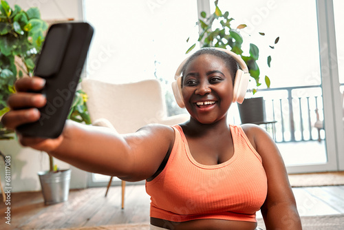 Gadgets for distance sport. Afro lady in wireless headset having video call on smartphone during daily workout at home. Happy curvy woman talking with trainer online for weight loss exercises.