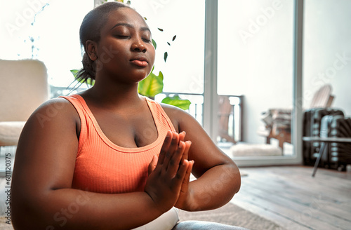 Relaxing yoga. Serene curvy woman in sport attire clasping hands in namaste and keeping eyes closed during meditation. African female sitting in lotus pose at living room and feeling peace of mind.