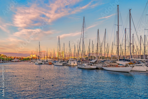 Yachts moored in the Port of Barcelona at sunset, Spain. Many boats with masts in the bay of the Mediterranean Sea against the backdrop of the city coastline illuminated by the rays of the setting sun