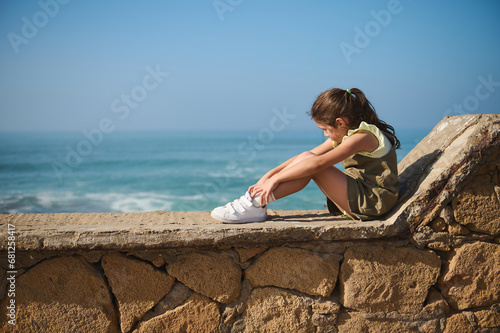 Full size portrait of a cute little kid girl sitting on a stone parapet and dreamily looking aside admiring Ocean storm