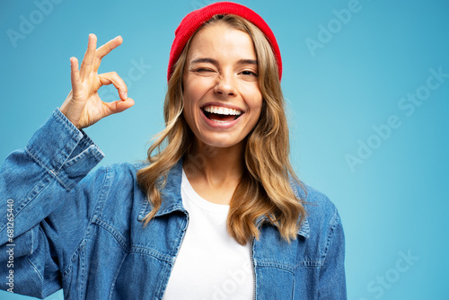 Smiling beautiful woman winking wearing stylish red hat showing ok sign isolated on blue background. Portrait happy modern hipster female with white teeth looking at camera in studio, dental concept 