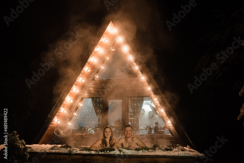 Man hug woman enjoying thermal spa in snowy forest. Couple relax in hot bath outdoors near house with garlands at night. Winter holidays in mountains, hot water treatments concept. Honeymoon vacation