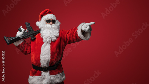 A man in a Santa Claus suit, wearing black sunglasses, poses with toy guns in hand against a red backdrop