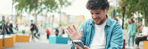 Clouseup, young bearded man in denim shirt sitting in wireless headphones making video call on mobile phone on busy street modern city background.
