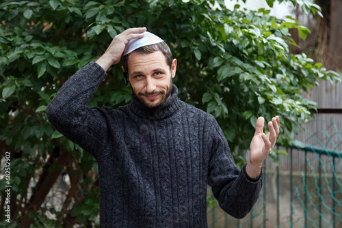 Religious Israeli caucasian man with smile face, holding a kippah, hand to his head. Handsome smiling bearded Jewish holding yarmulke for wind over his head while looking at camera. Looks surprised
