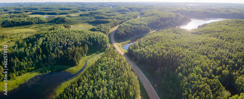 An aerial view of an extensive boreal forest with mixed tree types and scattered small lakes. A gravel road winds through the forest. The sun is reflecting off one of the lakes creating a sun glare on