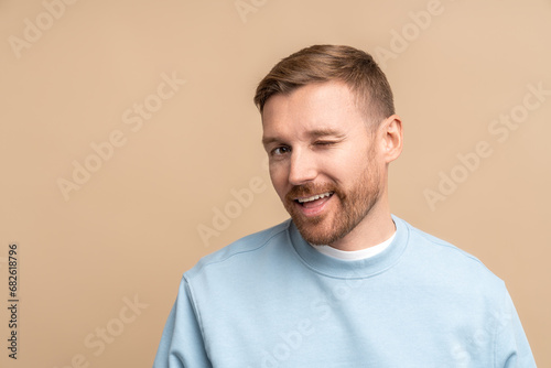 Flirting man winks look at camera, studio portrait isolated on beige. Bearded middle aged guy smiling winking playfully jokingly impishly with one eye, hinting, hidden intent, non-verbal communication