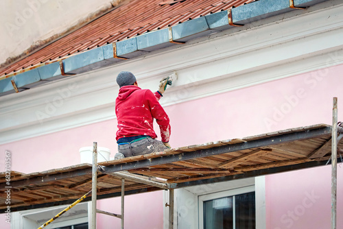 Worker on scaffolding paint facade wall of historic building. Utility worker paints building facade. Repair and restoration building facade. Man painting house wall with paintbrush
