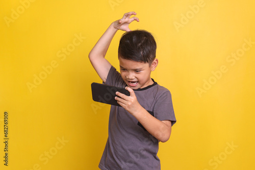 Angry little boy playing online game on smartphone isolated on yellow background