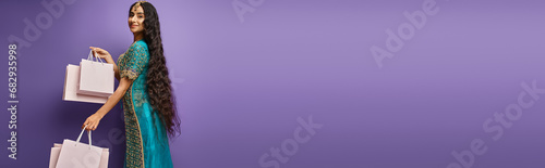 attractive jolly indian woman in blue sari posing with shopping bags on purple backdrop, banner