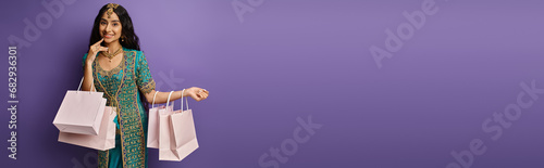 happy beautiful indian woman in blue sari posing with shopping bags on purple background, banner