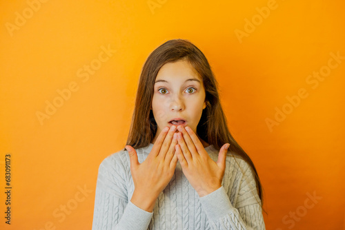 Shocked little girl with big dark brown eyes near the flame-colored background clothed in grey sweater is holding her hand over her mouth because of shock.