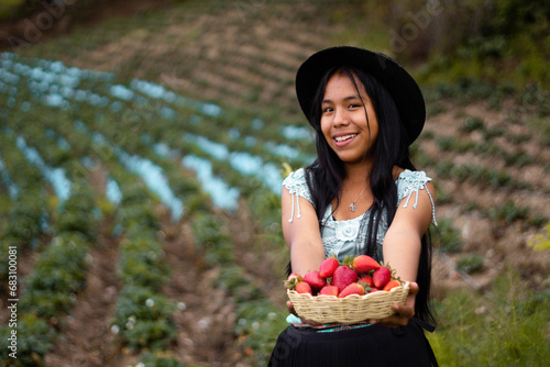 Una joven campesina con sombrero sosteniendo un tazón lleno de frutas y comiendo fresas maduras en el campo después de la cosecha