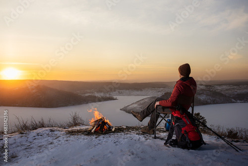  A man hiker sitting on the top of the mounting near campfire and enjoying yellow sunset at winter nature. Travel, lifestyle, freedom concept.