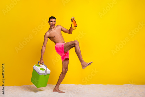 Full length photo of funny excited shirtless man holding refrigerator inviting drink beer empty space isolated yellow color background