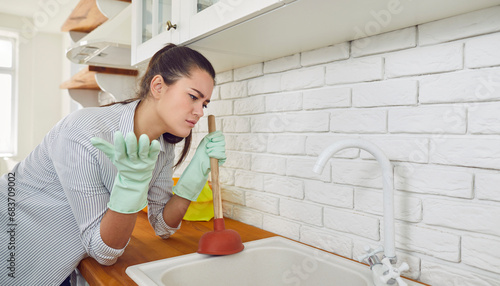 Young woman having a problem with the blocked kitchen sink. Sad girl doing work about the house and trying to unblock the clog in the kitchen sink drain with a plunger. Housework problem concept