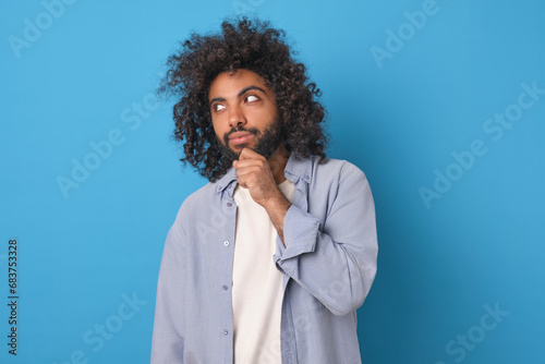 Young thinking curly Arabian man scratches chin and thinks about plan for next week or is trying to come up with idea for launching own money-making business stands posing in blue studio.