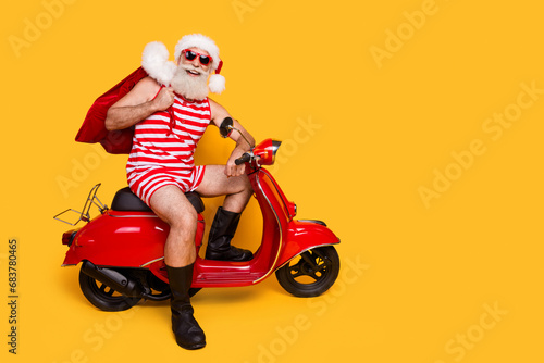 Full length photo of funny positive elderly guy wear new year swimsuit hat delivering presents empty space isolated yellow color background