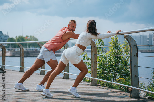 young cheerful couple trains outdoors, stretches their calf muscles on the embankment, leaning on fences. A male coach helps female to prepare for a sports girl's competition