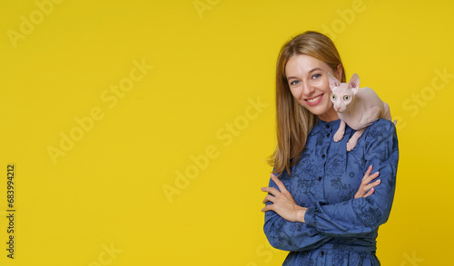 Smiling blond woman showcases unique bond with Egyptian cat Sphinx as it sits on shoulder against yellow background. Special companionship between woman and exotic feline friend, pet owner and cat.