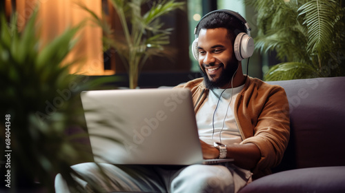 Young smiling afro male working on laptop. Man wearing headphones for listen music and sitting puffs.