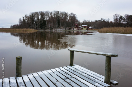 Scandinavian landscape with a lake in late autumn, Small wooden bridge sprinkled with fresh snow, Traditional Swedish red houses and a forest with bare trees reflected in calm water of Lake Mälaren