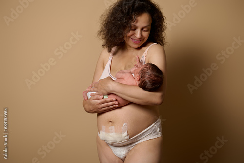 Authentic mom in underwear, smiles looking at her newborn baby she's holding, shows her body with postpartum scars flaws