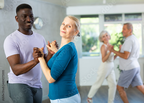 Cheerful senior woman practicing passionate samba with interested african american man in dance class for adults. Amateur dancing concept
