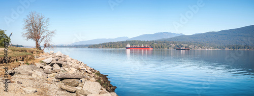 Cargo ship anchored in inlet in front of mountain scene. Summer marine transport panorama. Burrard Inlet, Inner Harbour, Vancouver, BC, Canada. View of North Shore Mountains. Selective focus.