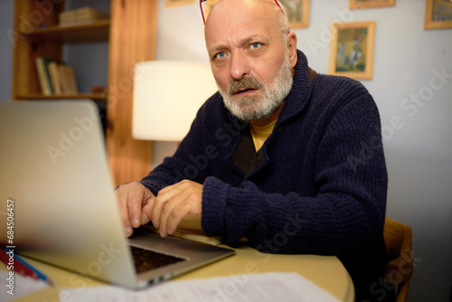 Side view of thoughtful senior novelist in elegant cardigan creating new plot of his novel, sitting in his cabinet at table in front of laptop, typing, looking at camera with thoughtful face
