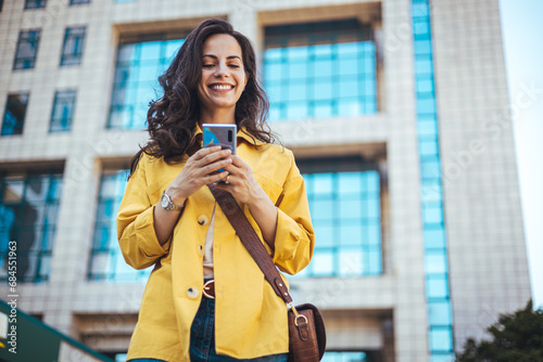 Concentrated female pedestrian in stylish outfit text messaging on mobile phone while walking along pavement near contemporary building. Focused woman strolling on sidewalk and reading message 