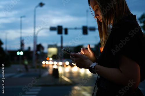 Close up shot of woman using smartphone on city street with bokeh lights at night. Mobile phone in female hands outdoors