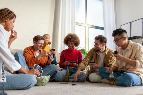 multiracial group of young people sitting at home with beer and popcorn and playing cards with friends
