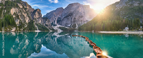 Panoramic photo of Lago di Braies, Pragser Wildsee in the Dolomites. View of the whole green-blue lake through wooden boats on the mountain peak and the setting sun.