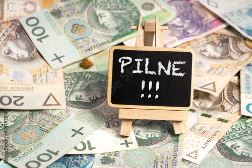 a small wooden writing board standing on scattered Polish zloty PLN banknotes, a chalk inscription "Pilne" on the black board, translation: Urgent (selective focus)