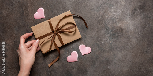 Female hand holds a brown envelope tied with a silk ribbon and pink silk hearts on a dark vintage background. Valentine's Day card. Top view, flat lay, copy space. Banner