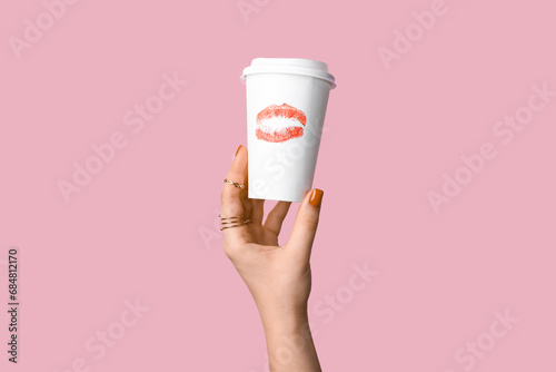 Female hand with red manicure holding paper cup of coffee on pink background